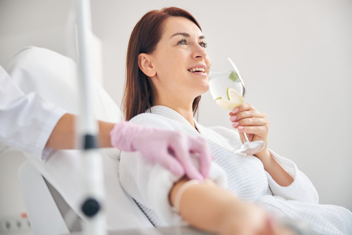 Joyous female patient drinking a healthy beverage while getting an IV Vitamin Therapy in a beauty clinic